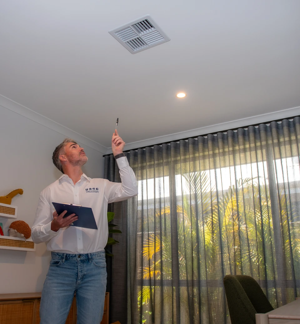 Maintenance worker with clipboard inspecting a ceiling vent in a bright living room.