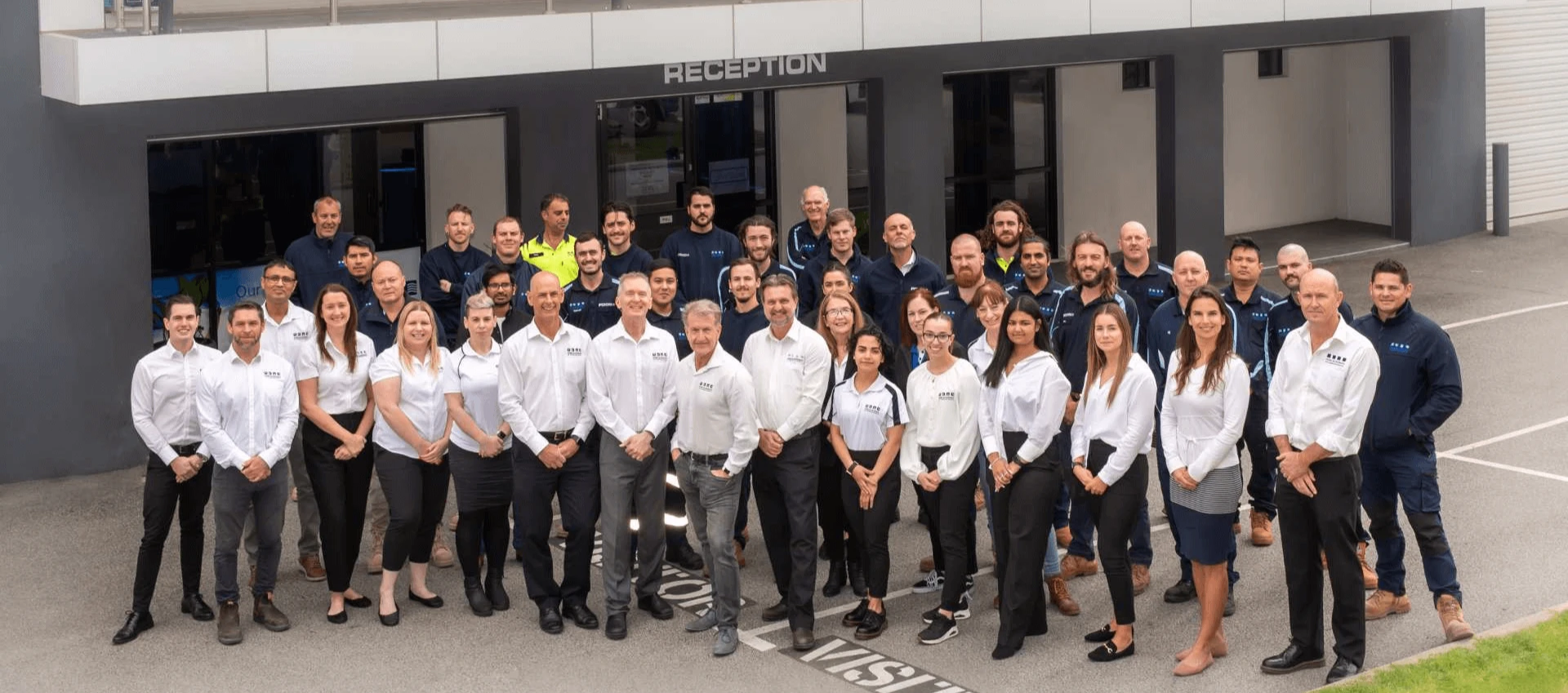 Group of employees in white shirts and dark uniforms posing outside a modern building’s reception area.