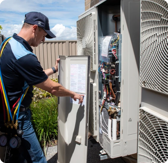 HVAC technician in a cap and tool belt inspects an outdoor air conditioner with the service panel open, exposing wiring and components.