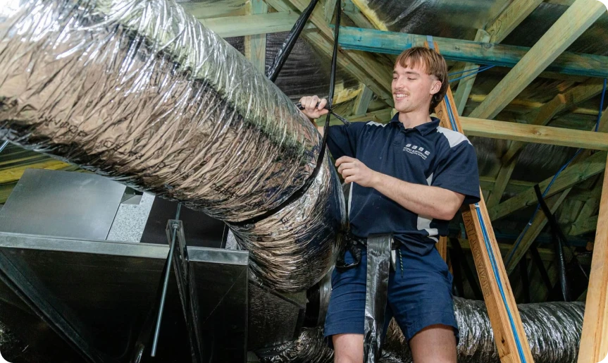 HVAC technician inspecting insulated air ducts in an attic while standing on a ladder Fielding the ducts with a tool, wearing a navy uniform.