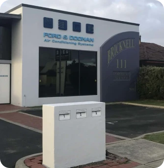Front exterior of an air‑conditioning service shop with blue signage on a white building; a white pedestal with three mail slots sits on a small brick-paved area in the foreground.