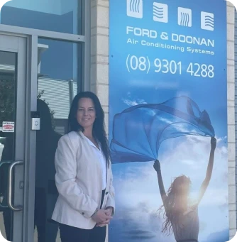 Woman in a light blazer stands at the storefront entrance beside a blue Ford & Doonan Air Conditioning Systems banner with a raised-arms figure and a phone number
