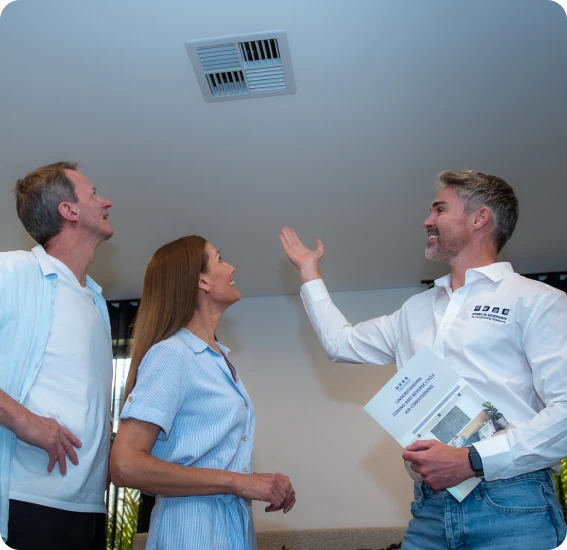 Three coworkers indoors look up at a ceiling vent as the man on the right gestures and holds a brochure.