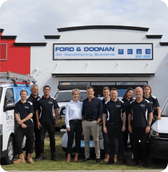 Group of Ford & Doonan air-conditioning technicians posing in front of a white building with the company sign and two vans outside.