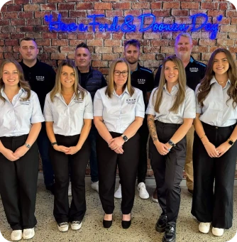 Group portrait of eight staff members in white shirts, standing in front of a brick wall with a blue neon sign.