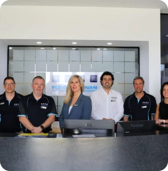 Six professionals standing behind a reception desk in a modern office, with a company logo visible on the frosted glass behind them.