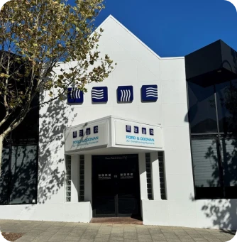 White modern office building with blue square logos above the entrance, and a tree casting shade.