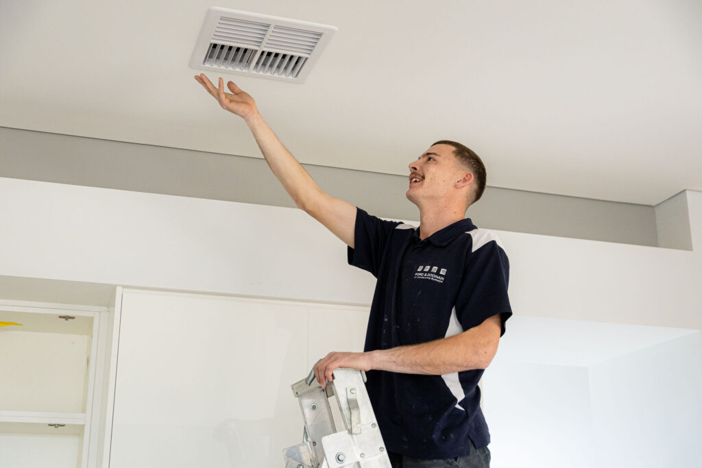 service-air-con - Ford & Doonan Air Conditioning Man on a ladder reaching a ceiling vent, installing or inspecting it in a bright room.